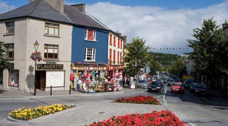 Een levendige straat in Westport aan de westkust van Ierland wordt gekenmerkt door kleurrijke winkelpuien zoals die van Flaherty en Thomas Moran, levendige bloemperken en wapperende vlaggen. Auto's en loofbomen omzomen de weg onder een halfbewolkte Ierse hemel. | Echt Ierland Vakanties