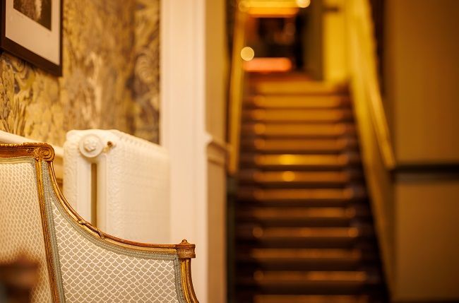 Een vintage gestoffeerde stoel met gouden rand staat bij een witte radiator in een warm verlichte hal van een charmant boetiek hotel in het centrum van Killarney; een trap met donkere houten treden en gouden verlichting gaat omhoog op de onscherpe achtergrond. | Echt Ierland Vakanties