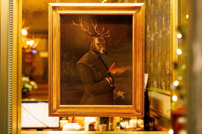 Een ingelijst schilderij van een hert in formele 18e-eeuwse kleding hangt aan een reflecterende muur en voegt eigenzinnigheid toe aan dit charmante boetiekhotel in het centrum van Killarney. Warme sfeerverlichting creëert een gezellige, elegante sfeer rond het portret. | Echt Ierland Vakanties