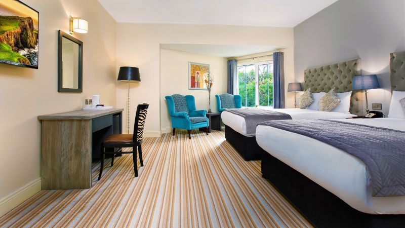 Een lichte hotelkamer in het Hillyard House Hotel in Castlewellan heeft twee grote bedden met grijze hoofdeinden, blauwgrijs beddengoed, groenblauwe fauteuils bij het raam, een gestreept tapijt, een bureaustoel met zebrastrepen, wandlampen, kunstwerken en een flatscreentelevisie. | Echt Ierland Vakanties