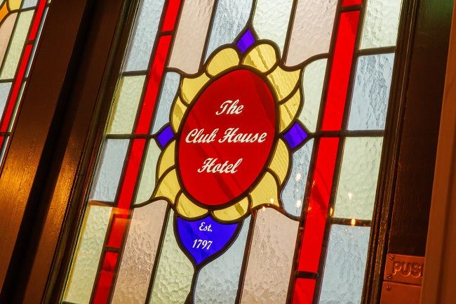 Glas-in-loodraam met in het midden een rode ovaal met de tekst "The Club House Hotel" in wit schrift, omringd door gele, blauwe en rode geometrische patronen - een charmant boetiekhotel dat doet denken aan die in het centrum van Killarney. | Echt Ierland Vakanties