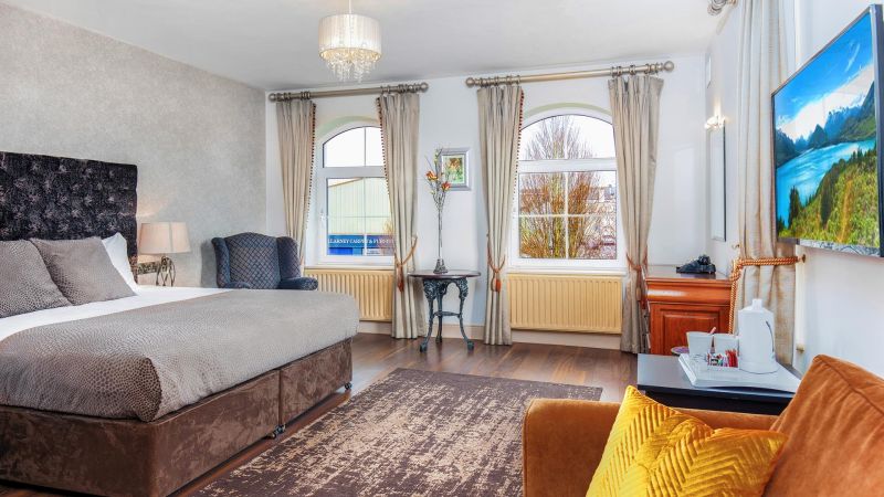 Een ruime, lichte slaapkamer in het Hillyard House Hotel in Castlewellan heeft een groot bed met grijs beddengoed, een gouden accentstoel, een donker vloerkleed en een stoel met wingback. Grote ramen met beige gordijnen vullen de kamer met natuurlijk licht. | Echt Ierland Vakanties