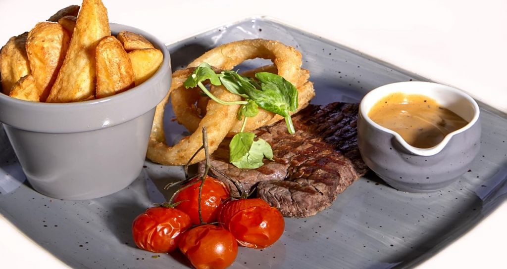 Op een grijs bord in het Brook Lodge Hotel in Killarney ligt een gegrilde steak, gouden uienringen met verse groenten, geroosterde trostomaten, dikgesneden friet in een kopje en een kleine grijze kan met romige bruine saus - alles netjes gerangschikt en goed verlicht. | Echt Ierland Vakanties