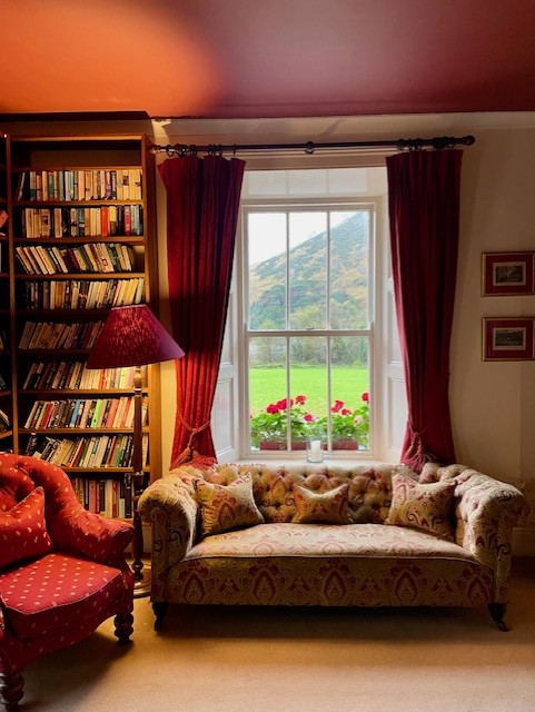 Een gezellige woonkamer, geïnspireerd op Boetiek hotel Westport, met een bank met patroon en een rode fauteuil. Een boekenplank staat vol met boeken, rode gordijnen omlijsten het raam en buiten liggen weelderig gras, rode bloemen en een schilderachtige berg. Een rode lampenkap maakt de look compleet. | Echt Ierland Vakanties