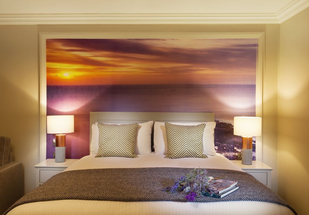 Een knus bed met twee kussens met zigzagpatroon en een grijze deken staat centraal onder een levendige muurschildering van een zonsondergang. Deze kamer, gevuld met oude charme, roept de intimiteit op van een boetiekhotel in het centrum van Kilkenny. | Echt Ierland Vakanties