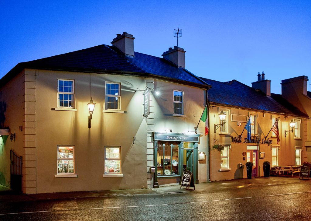 Een warm verlichte Ierse pub en winkel stralen in de schemering oude charme uit in het centrum van Kilkenny, met crèmekleurige muren, hangende lantaarns, Ierse vlaggen en een krijtbordbord bij de deur. De rustige natte straat gloeit onder een diepblauwe hemel. | Echt Ierland Vakanties