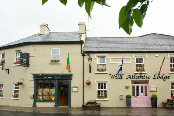 Een crèmekleurig gebouw met een leistenen dak straalt oude charme uit en huisvest de Wild Atlantic Lodge en Snug Bar. Roze deuren, vlaggen, zitbanken en bloembakken sieren de buitenkant en creëren een rustige sfeer die doet denken aan een boetiekhotel in het centrum van Kilkenny. | Echt Ierland Vakanties