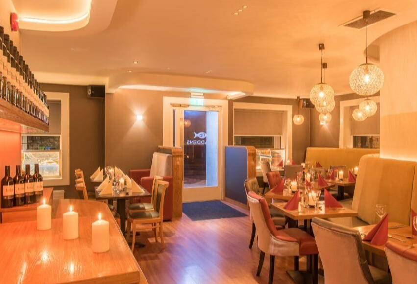 Een warm verlicht, modern restaurantinterieur met houten vloeren, beige en bruine stoelen, kaarsverlichte tafels met rode servetten en decoratieve bollampen. Het doet denken aan een gezellig hotel met eigen pub in Ballyvaughan, met wijnflessen op een plank bij de ingang. | Echt Ierland Vakanties