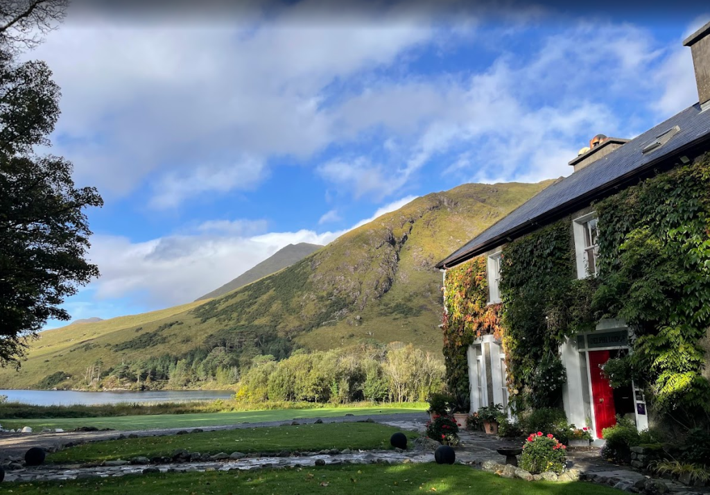 Een huis met met klimop bedekte muren en een felrode deur staat op de voorgrond, omringd door weelderig groen. Daarachter rijst een met gras begroeide berg op onder een blauwe hemel - een idyllisch landelijk tafereel dat doet denken aan een Boetiek hotel Westport escape. | Echt Ierland Vakanties