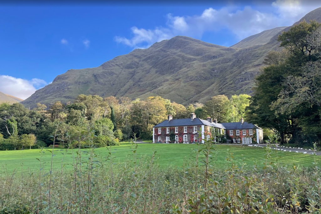 Een groot bakstenen landhuis met witte sierranden, dat doet denken aan Boetiek hotel Westport, ligt op een groen gazon omringd door hoge bomen. Op de achtergrond rijzen steile grasbergen op onder een blauwe hemel met verspreide wolken, terwijl struiken en planten de voorgrond vullen. | Echt Ierland Vakanties