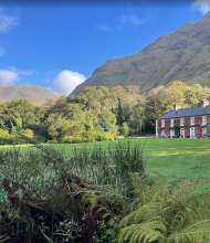 Een rood bakstenen landhuis met witte sierstrips staat naast een oprijlaan met gebogen grind, omringd door weelderige gazons en bomen. Achter het Boetiek hotel Westport rijzen zonovergoten heuvels op onder een blauwe hemel, terwijl varens en planten de voorgrond omlijsten. | Echt Ierland Vakanties
