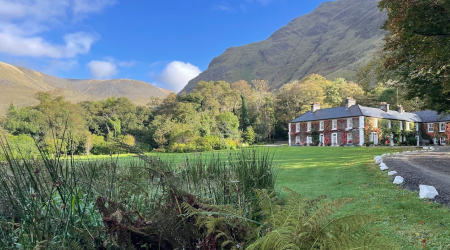 Een rood bakstenen landhuis met witte sierstrips staat naast een oprijlaan met gebogen grind, omringd door weelderige gazons en bomen. Achter het Boetiek hotel Westport rijzen zonovergoten heuvels op onder een blauwe hemel, terwijl varens en planten de voorgrond omlijsten. | Echt Ierland Vakanties