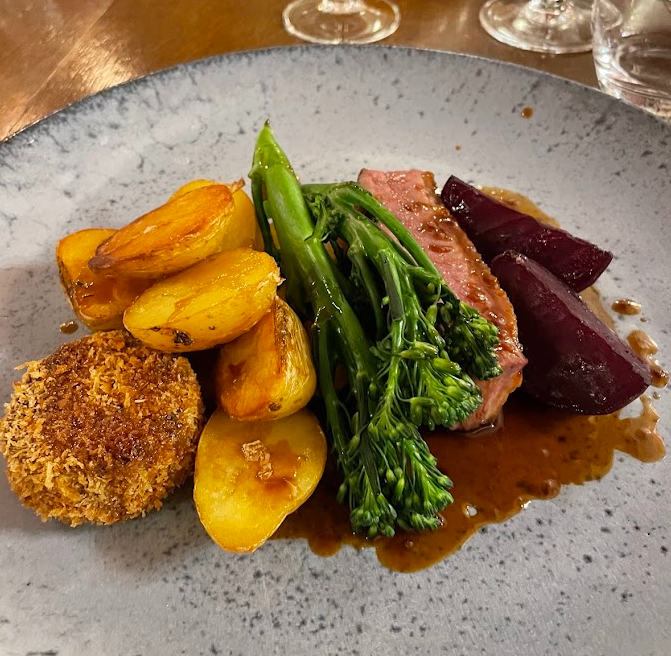 Een gastronomische maaltijd in Boetiek hotel Westport bestaat uit gesneden vlees met bruine saus, goudgeroosterde aardappelen, broccolini stengels, geglaceerde rode biet en een knapperige kroket, elegant geserveerd op een grijs gespikkeld bord met wijnglazen op de achtergrond. | Echt Ierland Vakanties