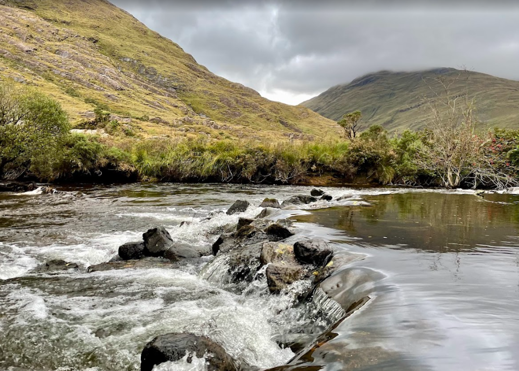 Een rotsachtig beekje stroomt door een groen, heuvelachtig landschap onder een bewolkte hemel bij Boetiek hotel Westport. Struiken en gras omzomen de waterkant, met mistige bergen op de achtergrond en kabbelend water over rotsen op de voorgrond. | Echt Ierland Vakanties