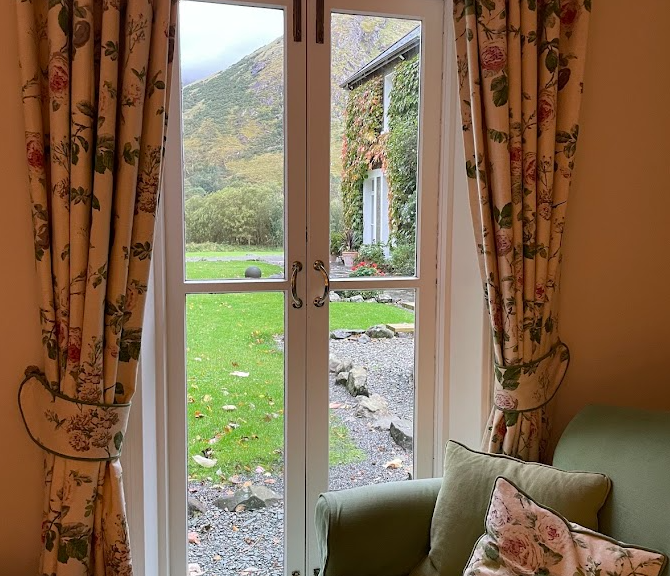 Een raam met gebloemde gordijnen opent naar een tuin met groen gras, een grindpad en een huismuur bedekt met klimop, die doet denken aan het serene uitzicht bij Boetiek hotel Westport. Op de voorgrond staat een fauteuil met een groen kussen. | Echt Ierland Vakanties