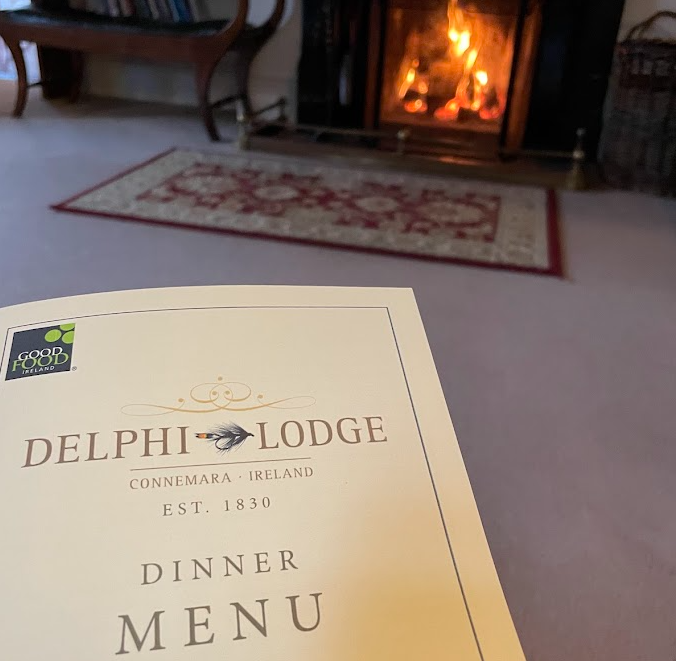 Op de voorgrond een dinermenu van Delphi Lodge, Connemara, Ierland, terwijl op de achtergrond een brandende open haard, een tapijt met patroon en een houten stoel de uitnodigende sfeer oproepen van een woonkamer in Westportstijl van een Boetiek hotel. | Echt Ierland Vakanties