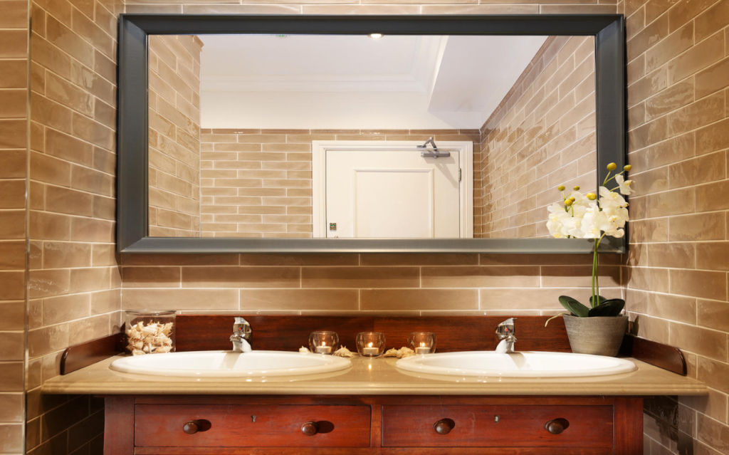 Een badkamer met bruine bakstenen muren straalt oude charme uit, met een grote rechthoekige spiegel boven een houten badmeubel met twee witte wastafels. Brandende kaarsen, schelpen in een glazen pot en een witte orchidee voegen elegantie toe die doet denken aan een boetiekhotel in het centrum van Kilkenny. | Echt Ierland Vakanties