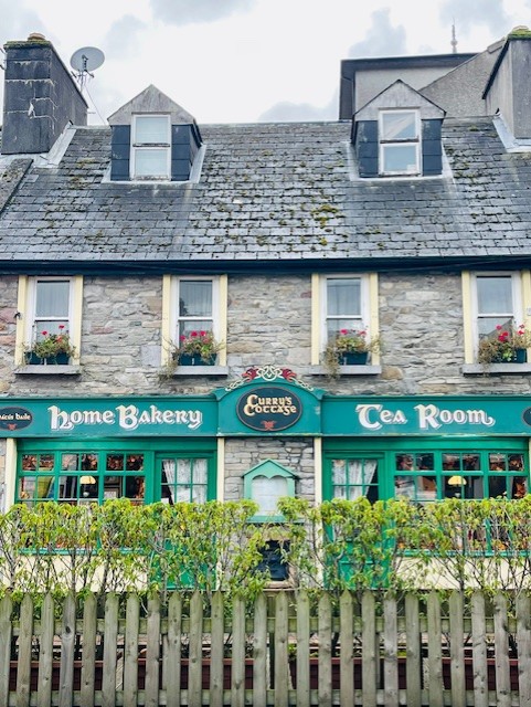 Een schilderachtig stenen gebouw met een leistenen dak huisvest Curran's Cookhouse, een thuisbakkerij en tearoom. Groen omrande ramen tonen bloemen, terwijl je vlakbij in Ballyvaughan een gezellig hotel met eigen pub vindt. De lucht is bewolkt. | Echt Ierland Vakanties