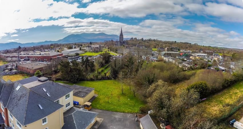 Een luchtfoto van Killarney toont verspreide gebouwen, een kerk met een hoge torenspits en het Brook Lodge Hotel te midden van groene velden, bomen en bergen onder een gedeeltelijk bewolkte hemel. Wegen kronkelen door het schilderachtige landschap. | Echt Ierland Vakanties