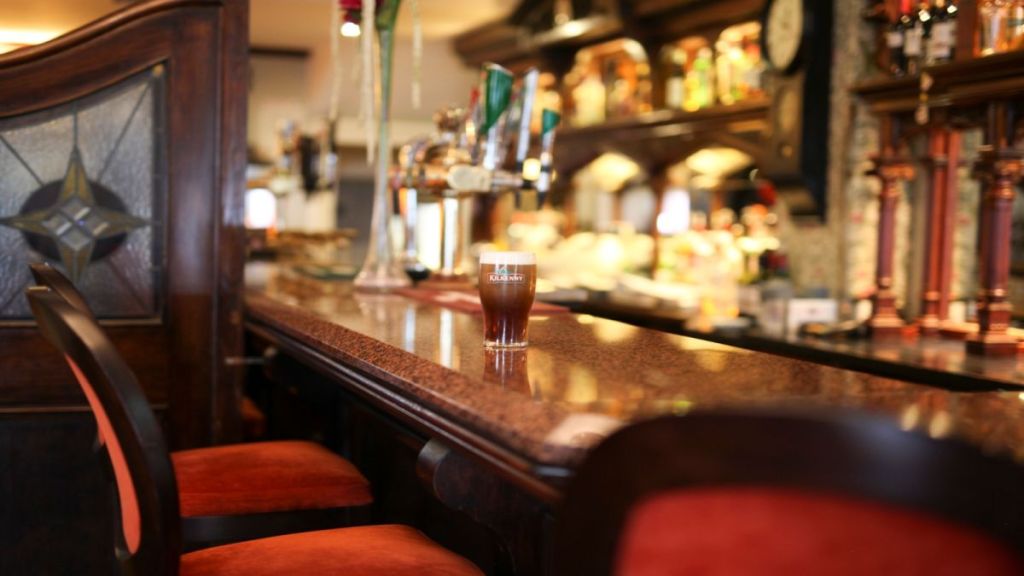 Een pint donker bier staat op een gepolijste, bruin marmeren bar in een warm verlichte pub in een charmant boetiekhotel in het centrum van Killarney, met houten lambrisering, rode stoelen en planken vol flessen en kranen op de wazige achtergrond. | Echt Ierland Vakanties