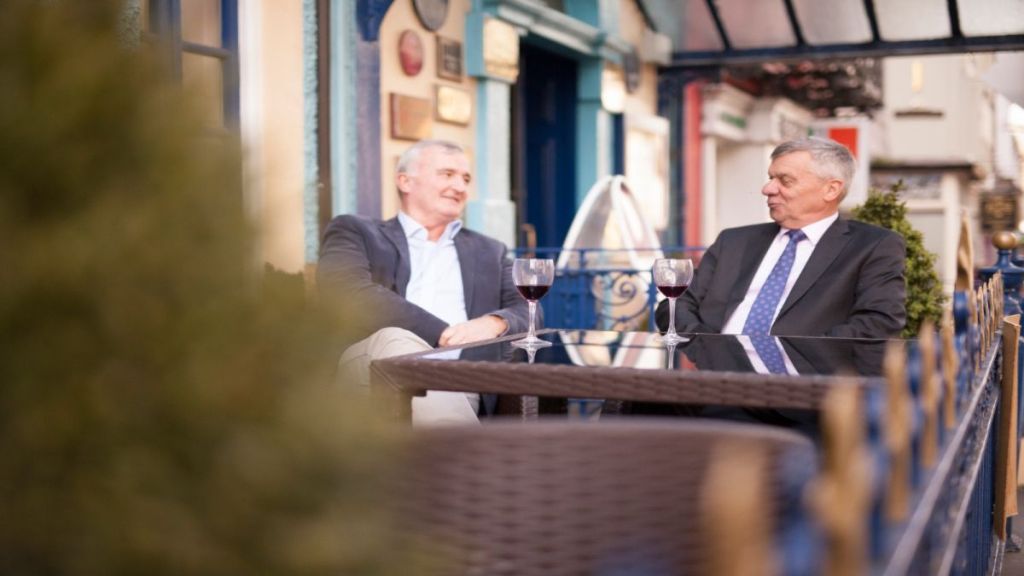 Twee oudere mannen in pak zitten aan een cafétafel in de buurt van een charmant boetiekhotel in het centrum van Killarney, elk met een glas rode wijn. Omringd door blauwe balustrades, potplanten en kleurrijke borden lijken ze in gesprek. | Echt Ierland Vakanties