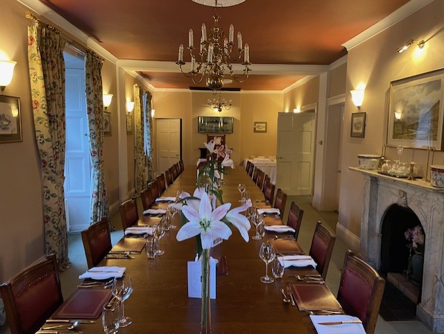 Een formele eetkamer in Boetiek hotel Westport heeft een lange houten tafel gedekt voor een maaltijd, met wijnglazen, borden en servetten. Witte lelies sieren het midden, terwijl kroonluchters, bloemige gordijnen en een open haard het klassieke decor compleet maken. | Echt Ierland Vakanties