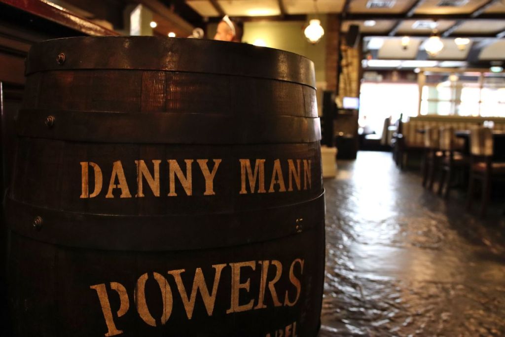Een groot houten vat met Danny Mann en Powers in gele letters staat in de rustieke, warm verlichte Killarney pub in Brook Lodge Hotel. Op de achtergrond staan lege houten stoelen en tafels, met zacht licht dat door de grote ramen naar binnen schijnt. | Echt Ierland Vakanties