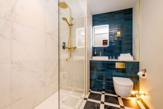 Een moderne badkamer, zoals die in een charmant boetiek hotel centrum Killarney, heeft een glazen douchewand, gouden armaturen, wit marmeren muren, blauwe accenttegels, een zwevend toilet, verlichte make-upspiegel en zwart-wit geblokte vloertegels. | Echt Ierland Vakanties