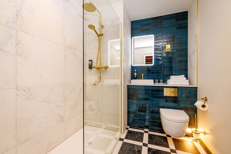Een moderne badkamer, zoals die in een charmant boetiek hotel centrum Killarney, heeft een glazen douchewand, gouden armaturen, wit marmeren muren, blauwe accenttegels, een zwevend toilet, verlichte make-upspiegel en zwart-wit geblokte vloertegels. | Echt Ierland Vakanties
