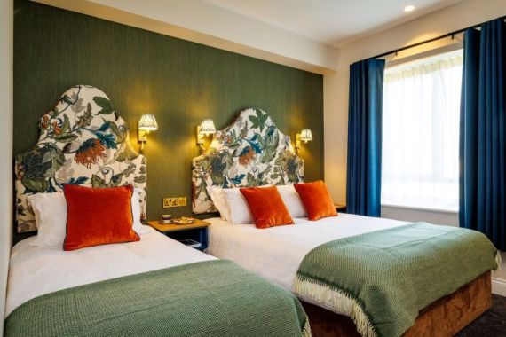 Een hotelkamer met twee bedden met wit beddengoed, groene spreien en oranje kussens. De bedden hebben hoofdeinden met bloemmotieven. Boven de bedden hangen twee wandlampen met gedessineerde kappen. De kamer heeft groene muren, blauwe gordijnen en een groot raam dat licht binnenlaat. | Echt Ierland Vakanties