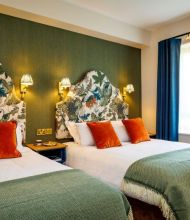 Een charmante boetiek hotelkamer in het centrum van Killarney met twee tweepersoonsbedden, witte lakens, groene spreien en oranje kussens. De kleurrijke bloemen hoofdeinden en blauwe gordijnen zorgen voor een frisse, lichte sfeer. | Echt Ierland Vakanties
