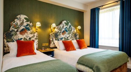 Een charmante boetiek hotelkamer in het centrum van Killarney met twee tweepersoonsbedden, witte lakens, groene spreien en oranje kussens. De kleurrijke bloemen hoofdeinden en blauwe gordijnen zorgen voor een frisse, lichte sfeer. | Echt Ierland Vakanties