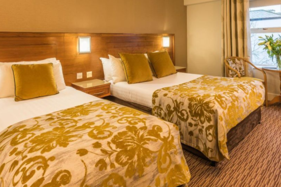 Een gezellige hotelkamer met twee tweepersoonsbedden, witte lakens, spreien met goudmotief en gouden kussens. Achter de bedden is een houten hoofdeinde met wandlampen. Dit gezellige hotel met eigen pub in Ballyvaughan biedt comfort en charme voor je verblijf. | Echt Ierland Vakanties