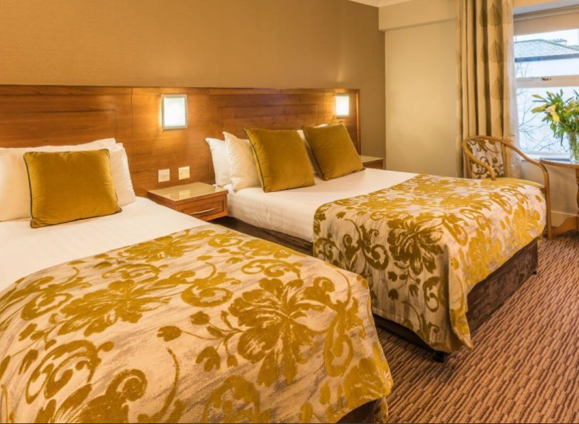 Een gezellige hotelkamer met twee tweepersoonsbedden, witte lakens, spreien met goudmotief en gouden kussens. Achter de bedden is een houten hoofdeinde met wandlampen. Dit gezellige hotel met eigen pub in Ballyvaughan biedt comfort en charme voor je verblijf. | Echt Ierland Vakanties
