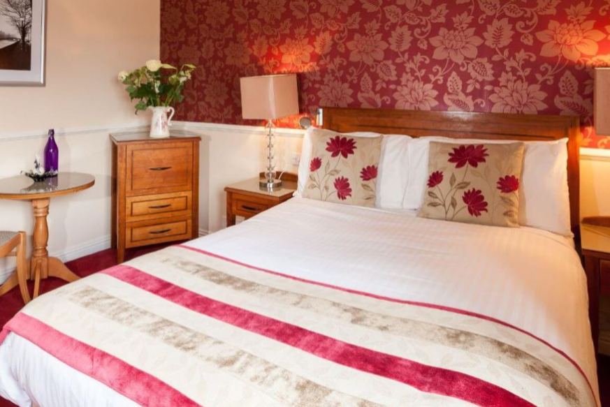 Een gezellige slaapkamer in een gezellig hotel met eigen pub in Ballyvaughan, met een tweepersoonsbed met wit linnengoed, een gestreepte beige en rode deken, gebloemde kussens, rood gebloemd behang, houten nachtkastjes met lampen en een vaas met bloemen. | Echt Ierland Vakanties