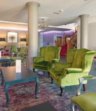 Een ruime, elegante lounge in het Brook Lodge Hotel in Killarney heeft groen en roze fluwelen fauteuils, donkere tafels en een kroonluchter. Lichte muren, kunst en een open haard sieren de kamer. Een trap met rood tapijt en paarse verlichting accentueren het decor. | Echt Ierland Vakanties