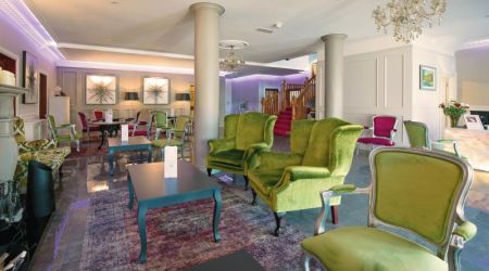 Een ruime, elegante lounge in het Brook Lodge Hotel in Killarney heeft groen en roze fluwelen fauteuils, donkere tafels en een kroonluchter. Lichte muren, kunst en een open haard sieren de kamer. Een trap met rood tapijt en paarse verlichting accentueren het decor. | Echt Ierland Vakanties