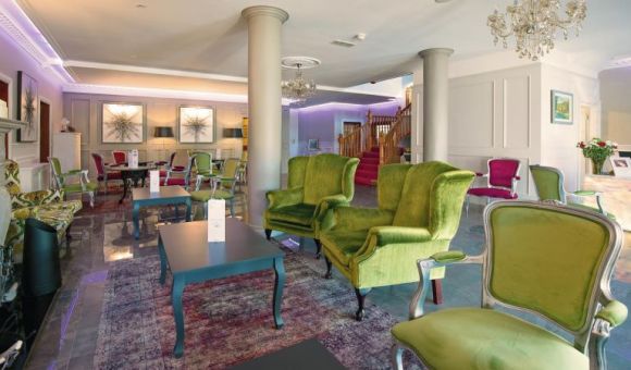 Een ruime, elegante lounge in het Brook Lodge Hotel in Killarney heeft groen en roze fluwelen fauteuils, donkere tafels en een kroonluchter. Lichte muren, kunst en een open haard sieren de kamer. Een trap met rood tapijt en paarse verlichting accentueren het decor. | Echt Ierland Vakanties