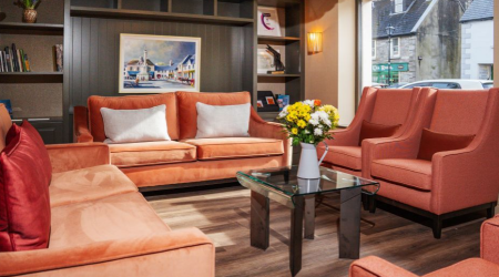Een gezellige lounge in een gezellig hotel met eigen pub in Ballyvaughan, met oranje banken en fauteuils rond een glazen salontafel met bloemen. Natuurlijk licht stroomt door een groot raam, terwijl boeken en ingelijste kunst de donkere muur erachter sieren. | Echt Ierland Vakanties