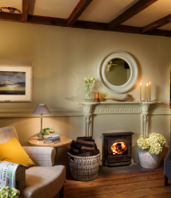 Een gezellige woonkamer met houten balken, een brandende houtkachel en twee fauteuils straalt oude charme uit. Zachte verlichting, kaarsen op de schoorsteenmantel en een raam met uitzicht doen denken aan een gastvrij boetiekhotel in het centrum van Kilkenny. | Echt Ierland Vakanties