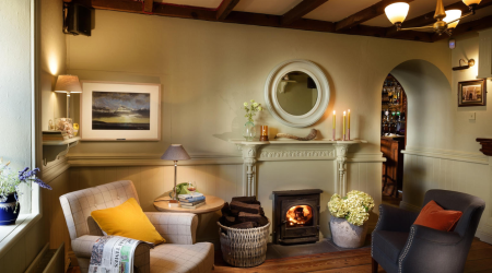 Een gezellige woonkamer met houten balken, een brandende houtkachel en twee fauteuils straalt oude charme uit. Zachte verlichting, kaarsen op de schoorsteenmantel en een raam met uitzicht doen denken aan een gastvrij boetiekhotel in het centrum van Kilkenny. | Echt Ierland Vakanties