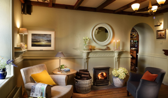 Een gezellige woonkamer met houten balken, een brandende houtkachel en twee fauteuils straalt oude charme uit. Zachte verlichting, kaarsen op de schoorsteenmantel en een raam met uitzicht doen denken aan een gastvrij boetiekhotel in het centrum van Kilkenny. | Echt Ierland Vakanties