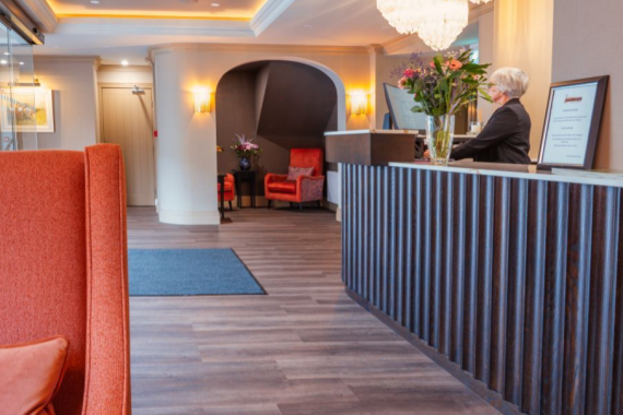 Een moderne hotellobby met houten vloeren, een receptie aan de rechterkant waar een persoon staat en een grote kroonluchter erboven. Dit gezellige hotel met eigen pub in Ballyvaughan heeft rode stoelen, bloemen en warme verlichting voor een gastvrije sfeer. | Echt Ierland Vakanties