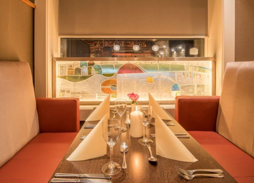 Een gezellig restaurant met rode kussens, een houten tafel voor vier personen met crèmekleurige servetten, glaswerk en bestek creëert een warme sfeer, net als een gezellig hotel met eigen pub in Ballyvaughan. Een roze roos siert het midden; glas-in-lood siert het raam. | Echt Ierland Vakanties