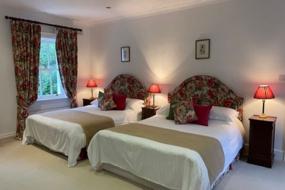 Een lichte slaapkamer in Boetiek hotel Westport met twee tweepersoonsbedden, elk met een gebloemd hoofdeinde, wit beddengoed, beige spreien en rode accentkussens. Bijpassende houten nachtkastjes met rode lampen en gebloemde gordijnen geven deze uitnodigende ruimte extra charme. | Echt Ierland Vakanties