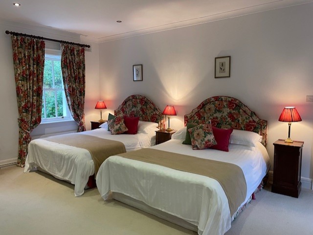 Een lichte slaapkamer in Boetiek hotel Westport met twee tweepersoonsbedden, elk met een gebloemd hoofdeinde, wit beddengoed, beige spreien en rode accentkussens. Bijpassende houten nachtkastjes met rode lampen en gebloemde gordijnen geven deze uitnodigende ruimte extra charme. | Echt Ierland Vakanties