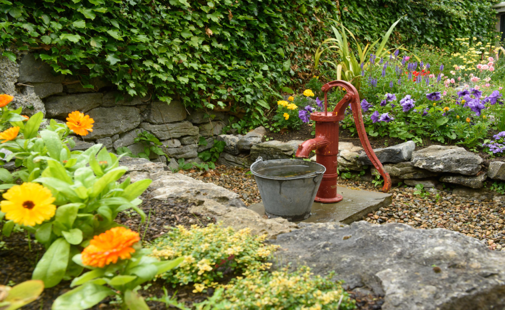Een rode handwaterpomp staat op een stenen plaat en straalt ouderwetse charme uit in een tuin met oranje, gele en paarse bloemen. Daarachter wijst een stenen muur bedekt met groene klimop op de elegantie van een boetiekhotel in het centrum van Kilkenny. | Echt Ierland Vakanties