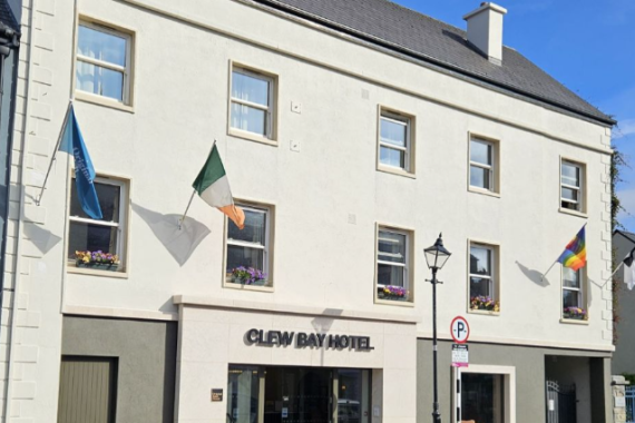 Een crèmekleurig gebouw van drie verdiepingen met "CLEW BAY HOTEL" boven glazen deuren. Drie vlaggen hangen boven ramen met bloemen. Het doet denken aan een gezellig hotel met eigen pub in Ballyvaughan, met een lantaarnpaal en een bord "Verboden te parkeren" onder een zonnige hemel. | Echt Ierland Vakanties