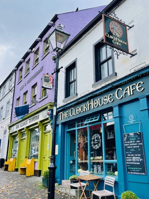 Een straatbeeld van The Clock House Café, blauw geschilderd met een krijtbordmenu en zitplaatsen buiten, staat naast The Clock Tavern-paars en groen, dat doet denken aan een gezellig hotel met eigen pub in Ballyvaughan, beide met klassieke uithangborden en vintage straatlantaarns. | Echt Ierland Vakanties