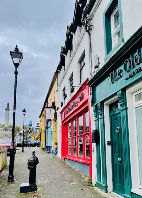 Een smalle straat met kleurrijke gebouwen, waaronder een pub met groene deuren en een rood café, ligt onder een bewolkte hemel. Vlakbij een zwarte lantaarnpaal en een waterpomp staat een gezellig hotel met eigen pub in Ballyvaughan. Vlaggen in de verte en een monument maken het kalme, enigszins natte tafereel compleet. | Echt Ierland Vakanties